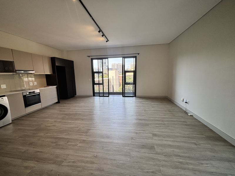 Spacious open-plan living area featuring modern kitchen cabinetry, integrated appliances, laminate flooring, and large sliding doors leading to a balcony with city views.