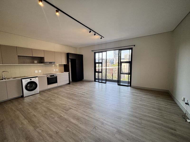 Modern open-plan living area featuring a sleek kitchen with built-in appliances, laminate flooring, and direct access to a private balcony through sliding glass doors.