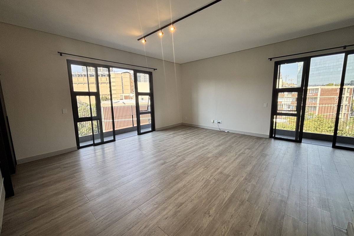 Spacious, modern living area featuring wood-look tile flooring, track lighting, and large black-framed glass doors leading to a balcony.