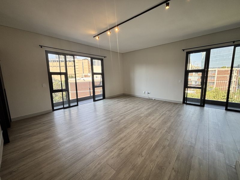Spacious, modern living area featuring wood-look tile flooring, track lighting, and large black-framed glass doors leading to a balcony.