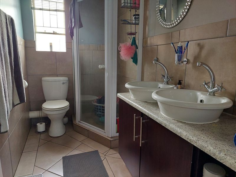 A functional bathroom featuring a double vanity with granite countertop, a shower enclosure, and a toilet, finished with neutral wall and floor tiles.