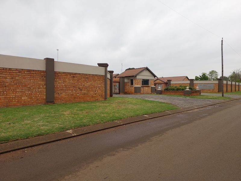 Street view of the townhouse complex entrance, featuring a secure gatehouse, brick perimeter walling, and paved access driveway.