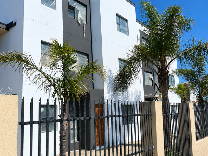 Modern apartment building exterior featuring a clean white and grey facade, secured by a wrought iron perimeter fence and masonry boundary wall.