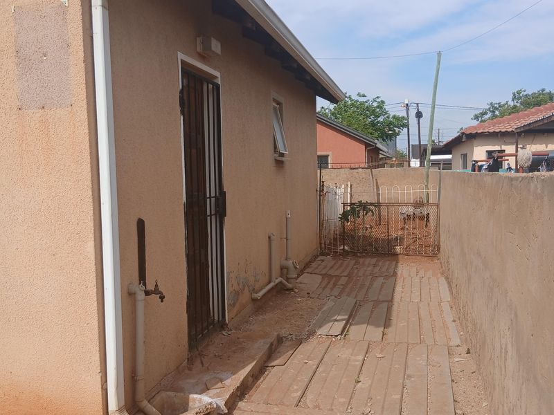 Side exterior view of the property featuring a concrete pathway, boundary wall, and external utility access with security gate.