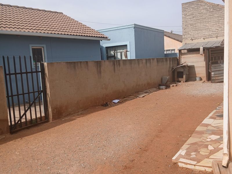 Rear exterior area featuring a boundary wall, metal security gate, and a small tiled patio section with unpaved ground.