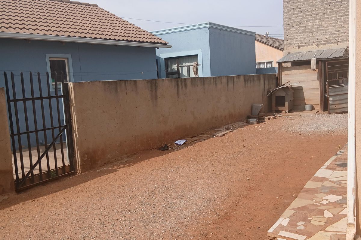 Rear exterior area featuring a boundary wall, metal security gate, and a small tiled patio section with unpaved ground.