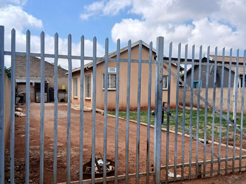 Front view of a single-story house featuring a steel palisade fence, gated entrance, and an unpaved driveway area.