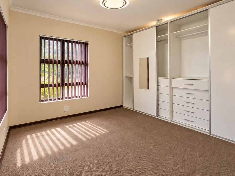 A clean, empty bedroom featuring neutral walls, carpeted flooring, and a large built-in wardrobe with integrated shelving and drawers.