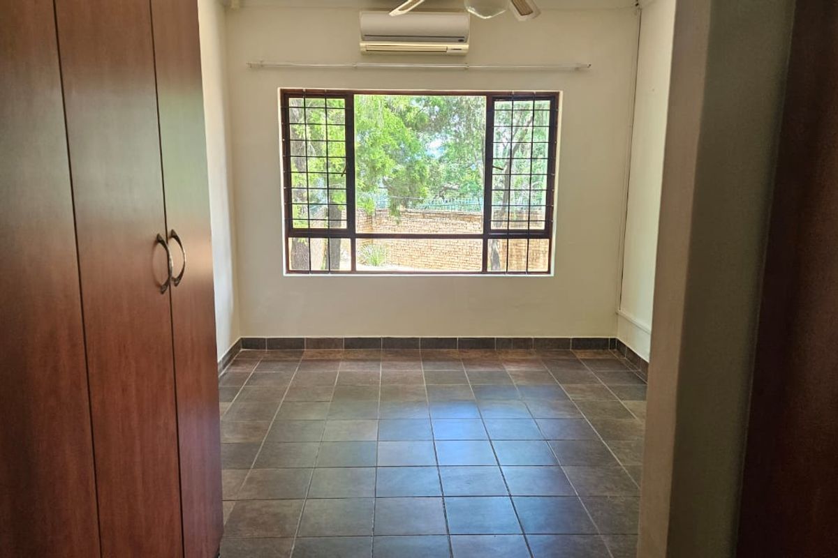 A clean, empty bedroom featuring tiled flooring, built-in wooden cupboards, a wall-mounted air conditioning unit, and a window with security bars.