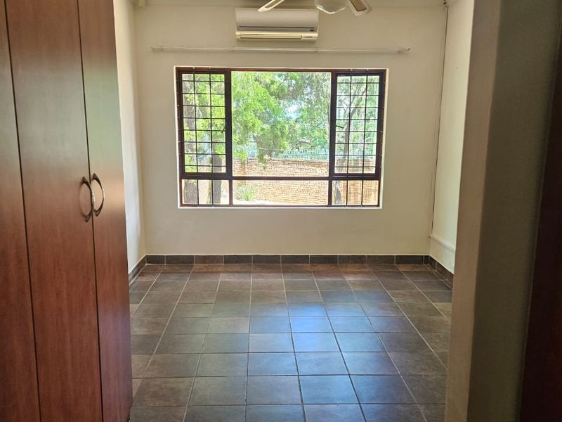 A clean, empty bedroom featuring tiled flooring, built-in wooden cupboards, a wall-mounted air conditioning unit, and a window with security bars.
