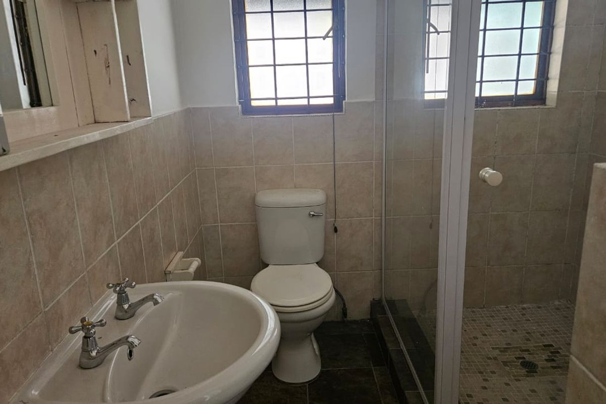 A functional bathroom featuring a pedestal basin, toilet, and a glass-enclosed shower with tiled finishes and security-barred windows.