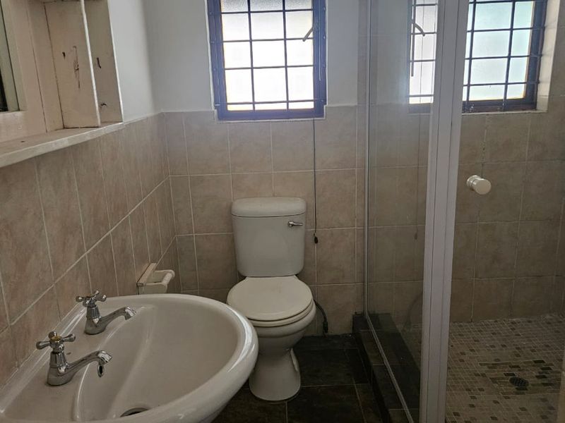 A functional bathroom featuring a pedestal basin, toilet, and a glass-enclosed shower with tiled finishes and security-barred windows.