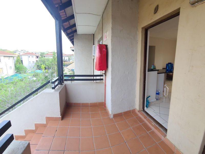 A covered balcony featuring tiled flooring and a metal balustrade, providing an outdoor space with views of the surrounding apartment complex.