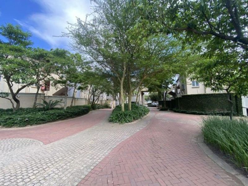 A well-maintained paved driveway entrance to the apartment complex, featuring mature trees and landscaped garden beds.