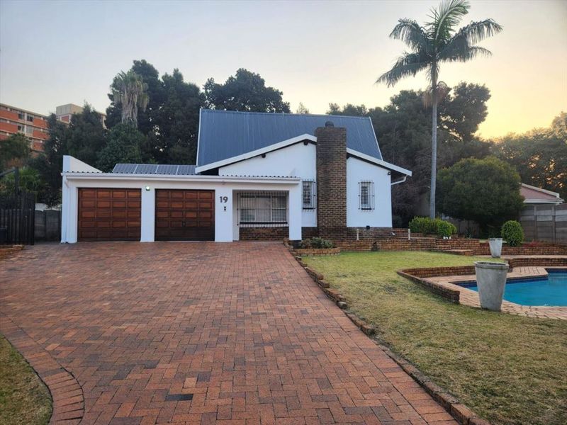 Front view of a well-maintained house featuring a double garage, brick driveway, swimming pool, and solar panels on a metal roof.