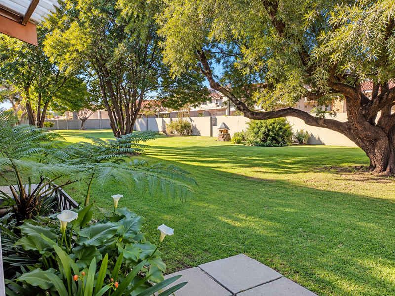 A spacious, well-maintained private garden featuring a lush lawn, mature trees, and a paved patio area, enclosed by a secure boundary wall.