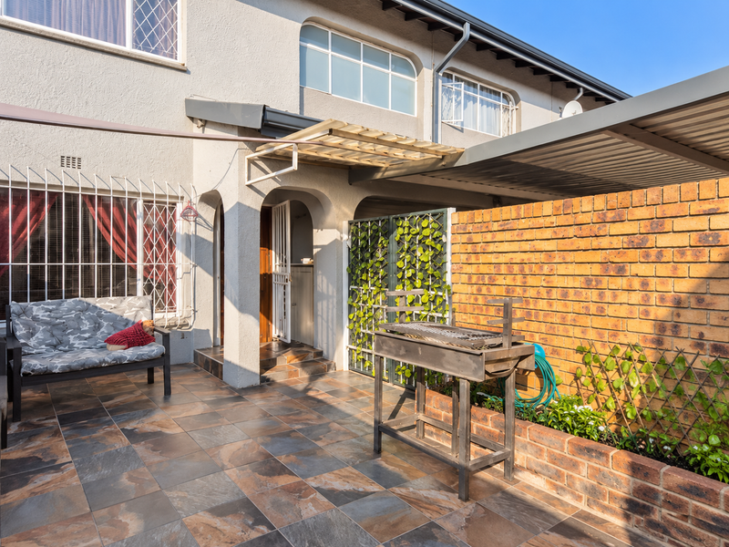 A paved outdoor patio area featuring a brick boundary wall, raised garden bed, and metal roof awning, providing a functional space for outdoor living.