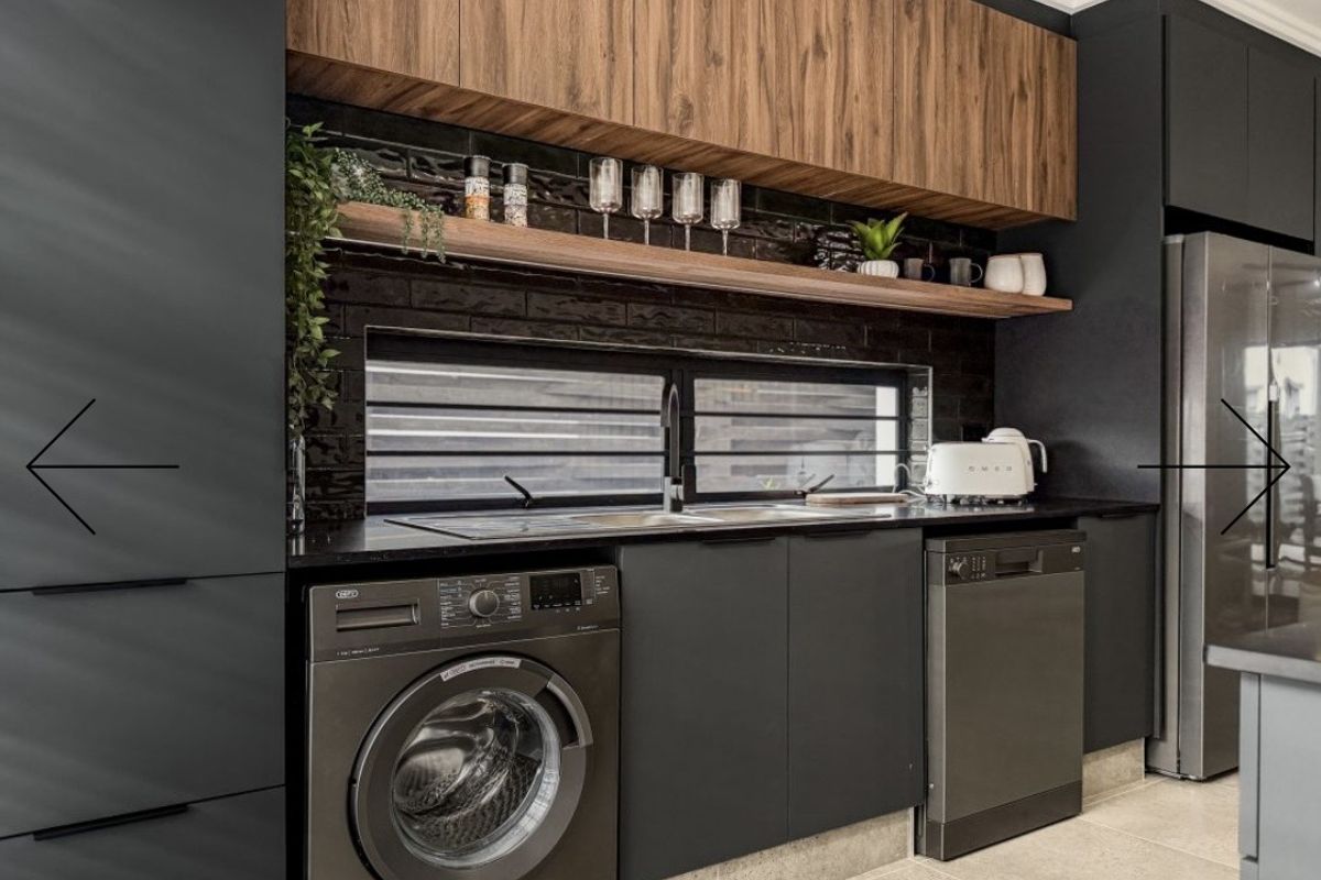 Modern laundry area featuring sleek dark cabinetry, timber shelving, a tiled splashback, and integrated appliances including a washing machine and dishwasher.