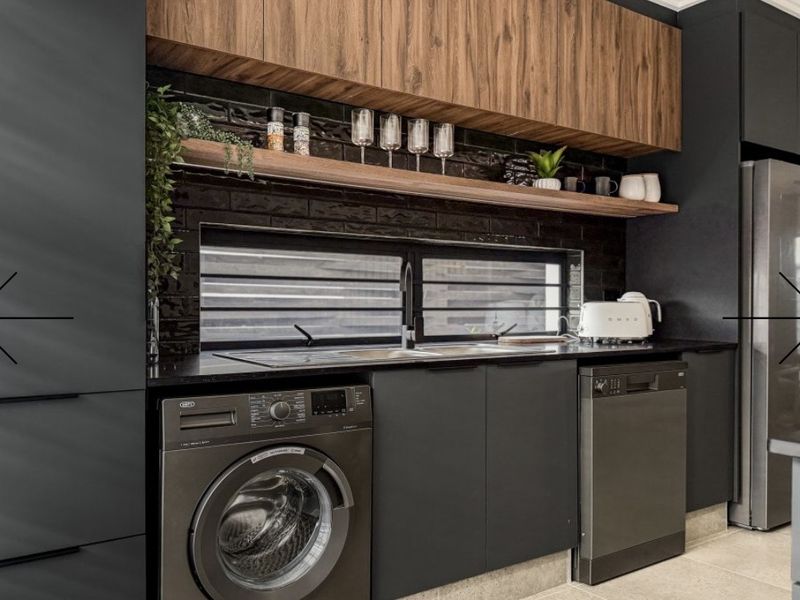 Modern laundry area featuring sleek dark cabinetry, timber shelving, a tiled splashback, and integrated appliances including a washing machine and dishwasher.