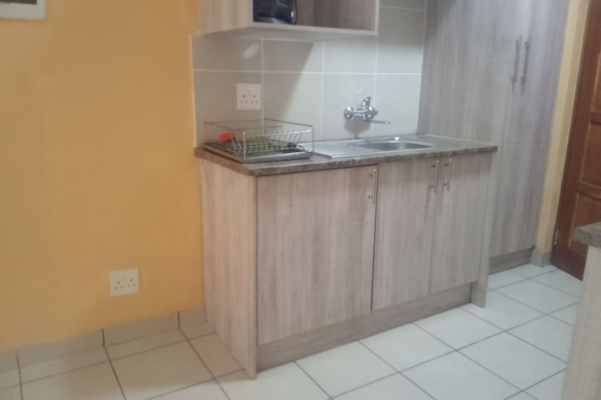 A compact kitchen area featuring light wood-grain cabinetry, a stainless steel sink, tiled backsplash, and neutral floor tiles.