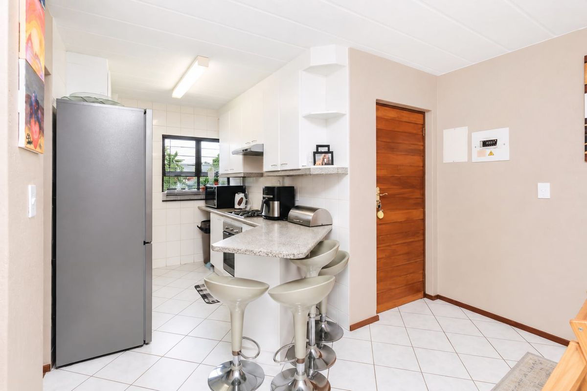 Compact kitchen featuring white cabinetry, stone countertops, a breakfast bar, built-in oven, and hob, with tiled flooring and a timber entrance door.