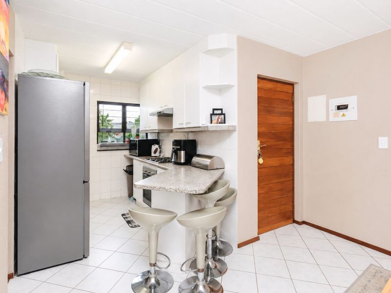 Compact kitchen featuring white cabinetry, stone countertops, a breakfast bar, built-in oven, and hob, with tiled flooring and a timber entrance door.