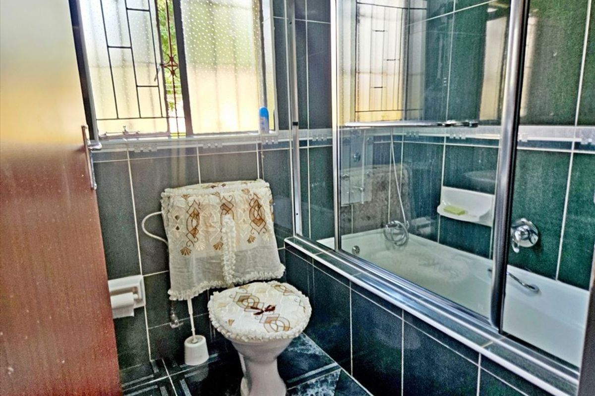 A compact bathroom featuring dark green wall and floor tiling, a built-in bathtub with a glass shower screen, and a window with security bars.