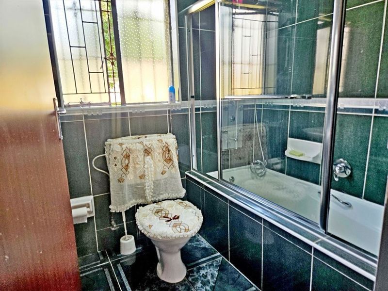 A compact bathroom featuring dark green wall and floor tiling, a built-in bathtub with a glass shower screen, and a window with security bars.