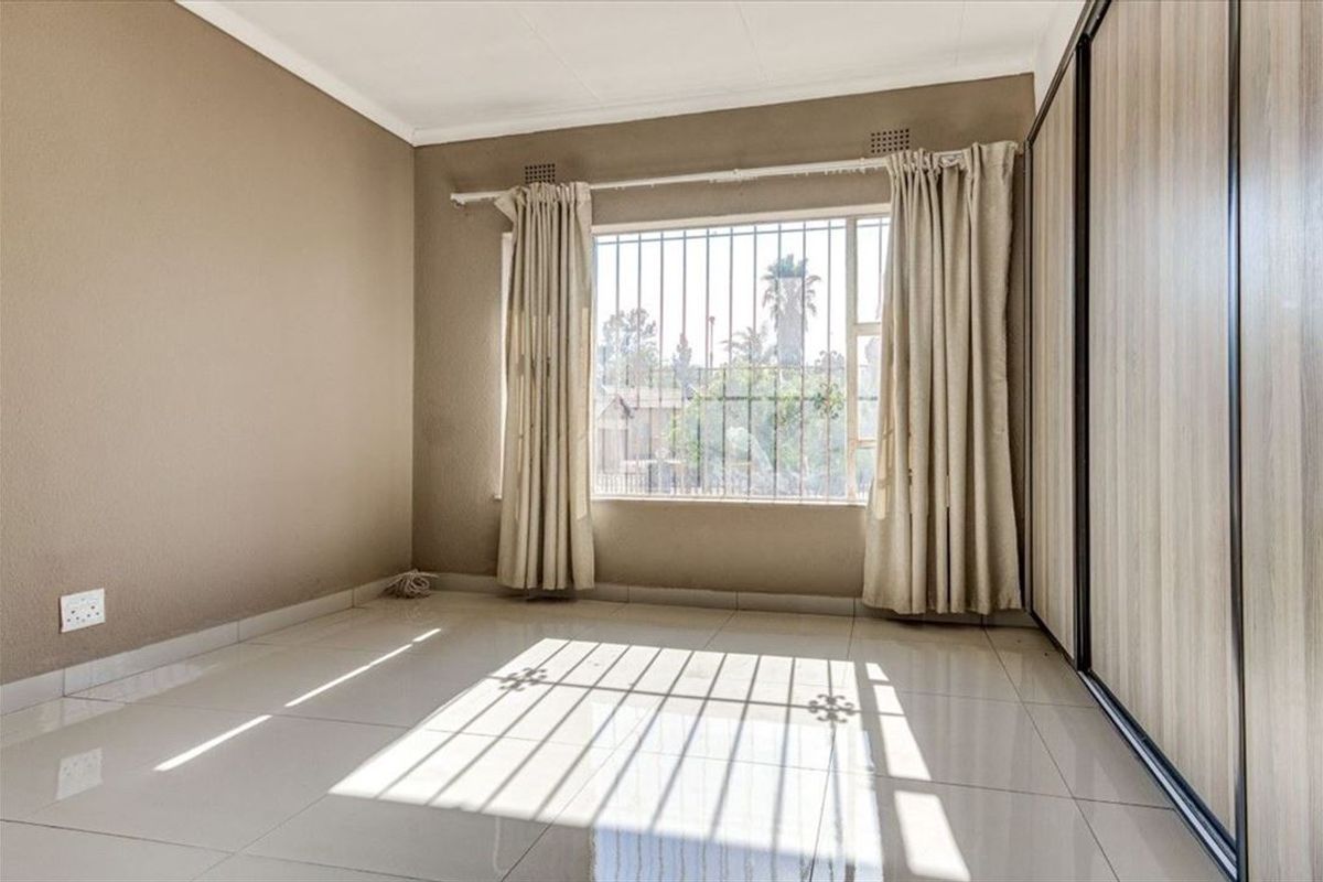 A bright, empty bedroom featuring neutral tiled flooring, built-in wooden cupboards, and a window fitted with security bars.