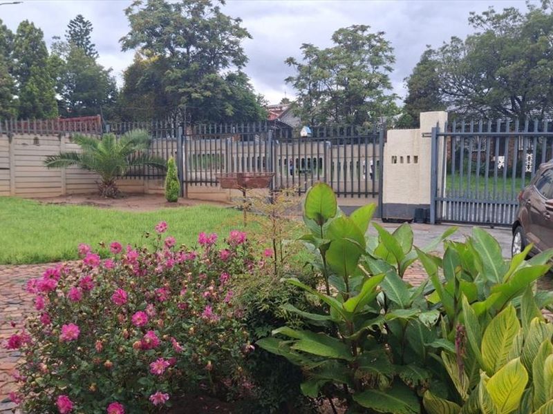 Front garden area featuring a brick paved driveway, established landscaping, and secure metal perimeter fencing with a gated entrance.