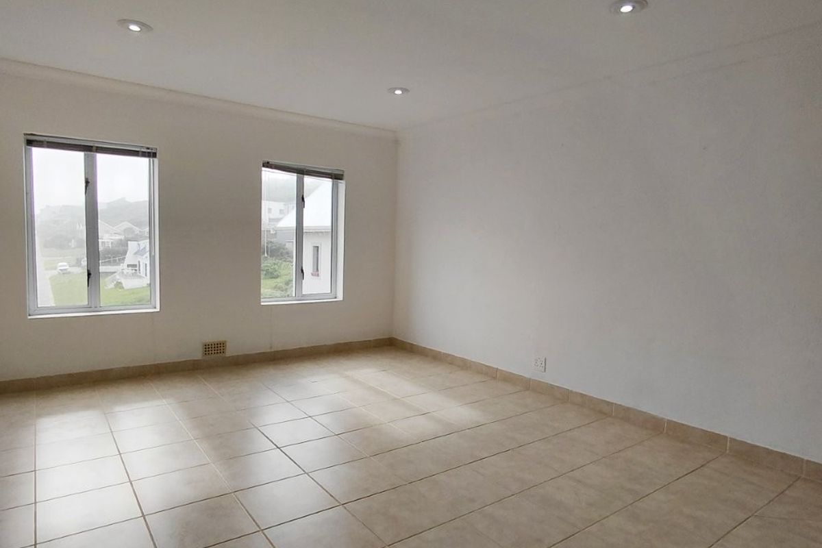 A bright, empty living room featuring neutral tiled flooring, recessed downlights, and large windows providing natural light.