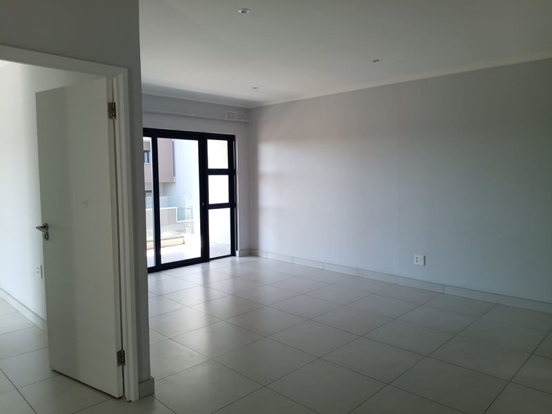 A bright, empty living room featuring neutral tiled flooring, recessed downlighting, and a large sliding glass door leading to an outdoor area.