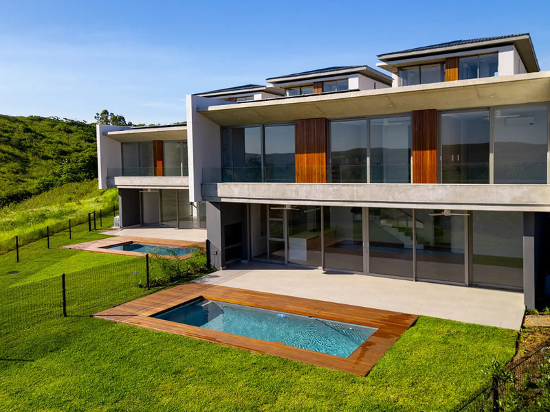 Modern multi-level residence featuring expansive glass sliding doors, timber accents, concrete balconies, and private swimming pools set within a landscaped garden.
