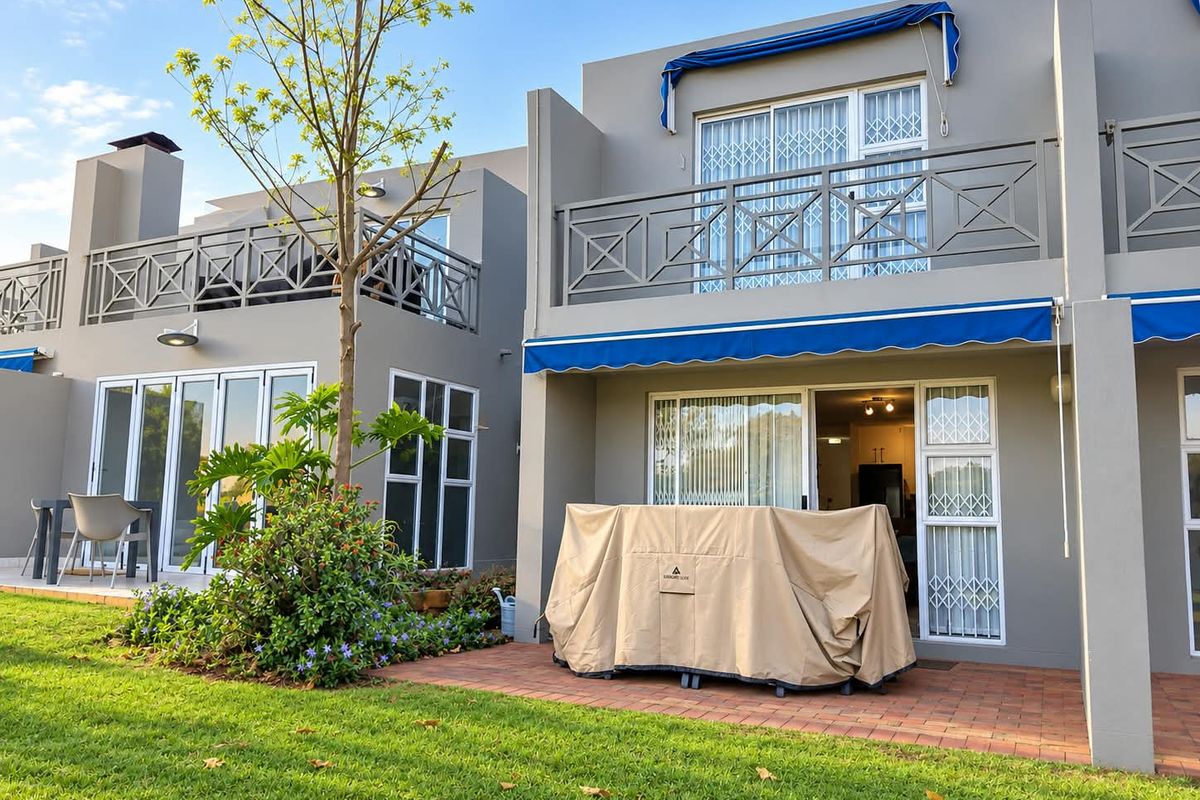 Rear view of the townhouse featuring a brick-paved patio, upper-level balcony with metal railings, and retractable awnings overlooking a landscaped garden area.