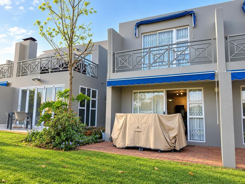 Rear view of the townhouse featuring a brick-paved patio, upper-level balcony with metal railings, and retractable awnings overlooking a landscaped garden area.