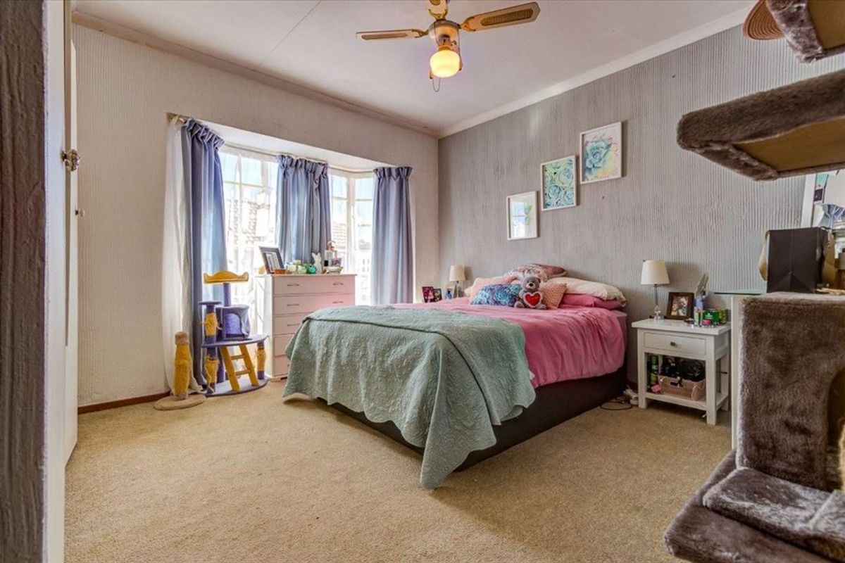 A bedroom featuring a bay window, ceiling fan, and textured wallpaper, providing a functional space with neutral carpet flooring.