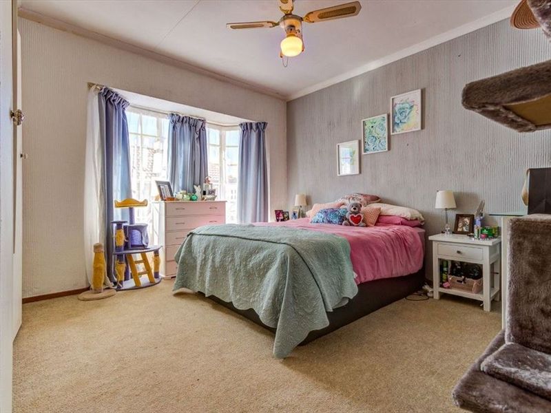 A bedroom featuring a bay window, ceiling fan, and textured wallpaper, providing a functional space with neutral carpet flooring.