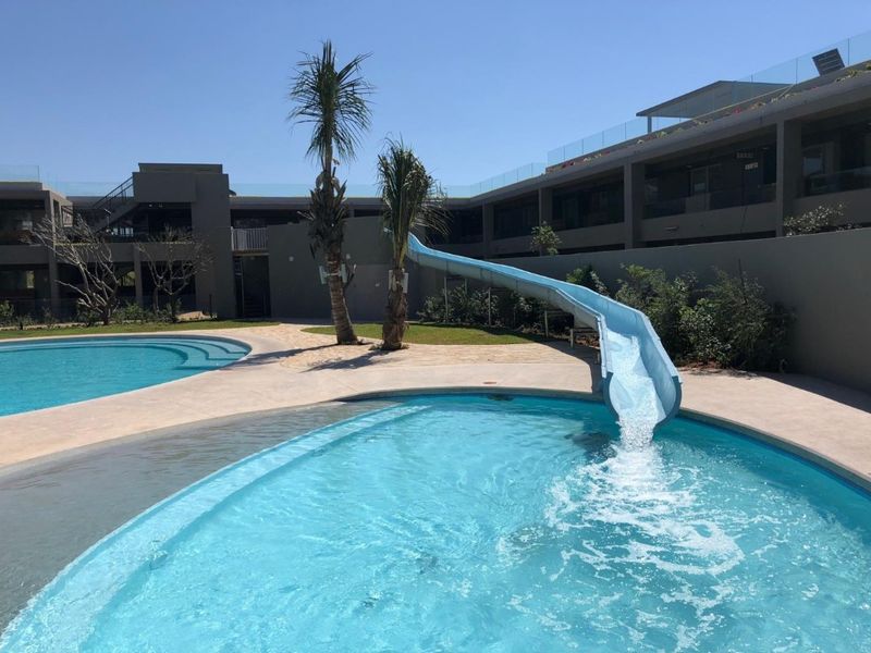 Communal swimming pool area featuring a water slide, concrete decking, and modern apartment building architecture with glass balustrades.
