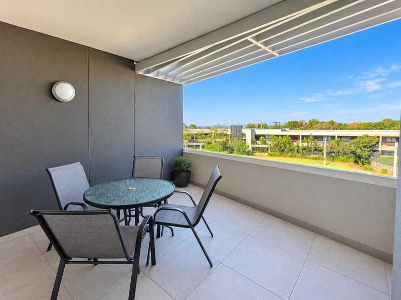 A modern, covered balcony featuring tiled flooring, a sleek overhead pergola, and a balustrade with pleasant views of the surrounding landscape.