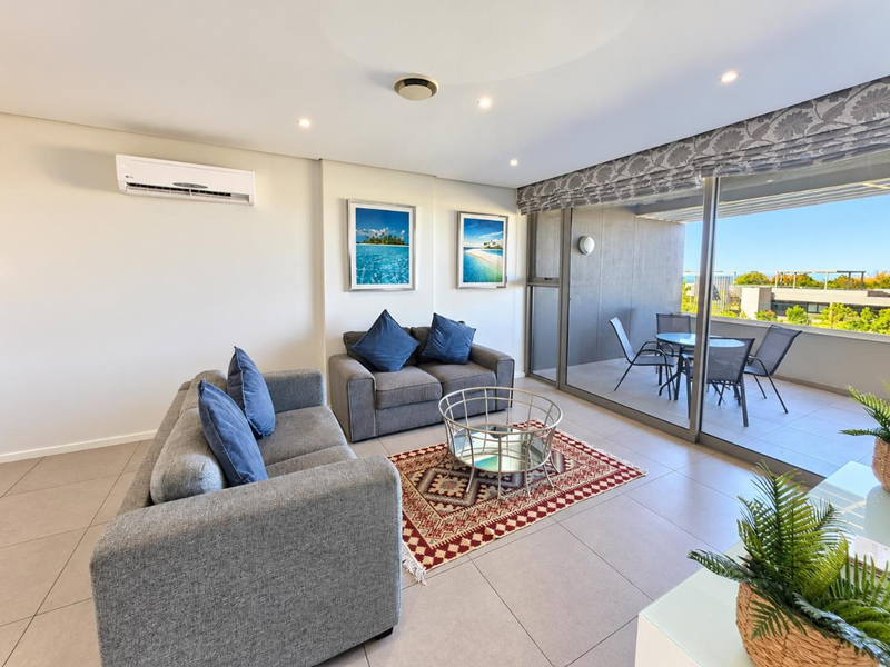 A bright, open-plan living area featuring tiled floors, recessed lighting, a wall-mounted air conditioner, and large sliding doors leading to a private balcony.