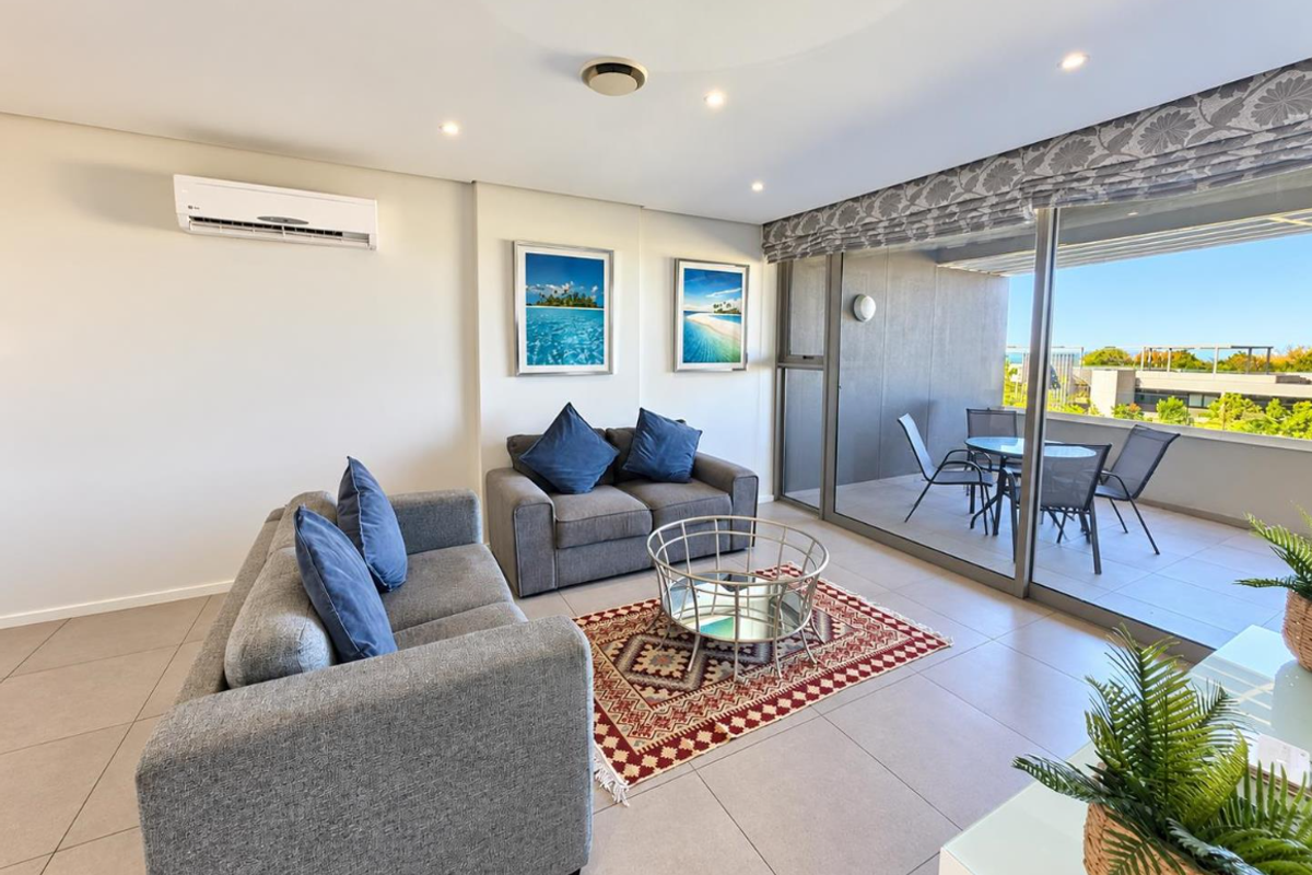 A bright, open-plan living area featuring tiled floors, recessed lighting, a wall-mounted air conditioner, and large sliding doors leading to a private balcony.