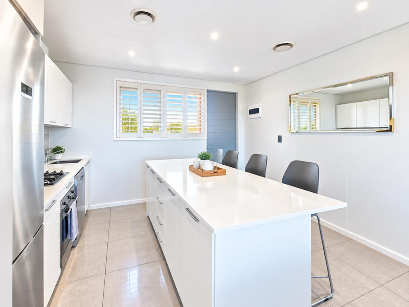 Modern kitchen featuring a large central island with stone countertops, integrated appliances, sleek white cabinetry, and neutral tiled flooring.