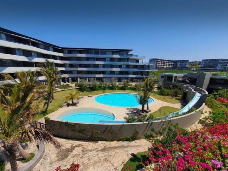 A well-maintained communal pool area featuring two circular swimming pools, a water slide, and landscaped gardens within a modern apartment complex.