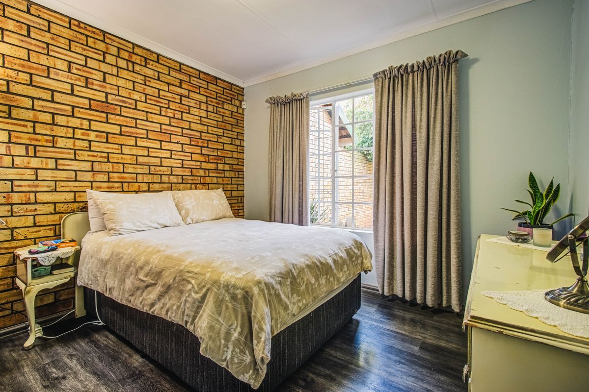 A cozy bedroom featuring an exposed brick accent wall, laminate flooring, and a window with security bars, offering a warm and textured aesthetic.