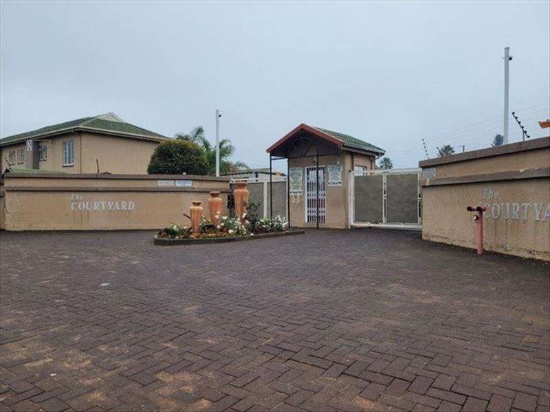 The entrance to The Courtyard complex featuring a paved driveway, security gate, and guard house with perimeter wall and electric fencing.