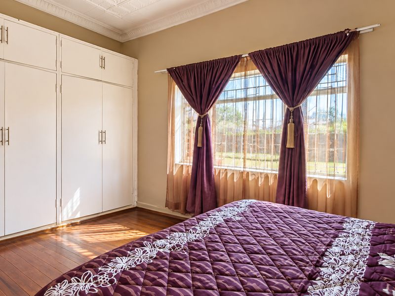 A well-maintained bedroom featuring extensive built-in cupboards, polished hardwood flooring, and large windows with security bars.
