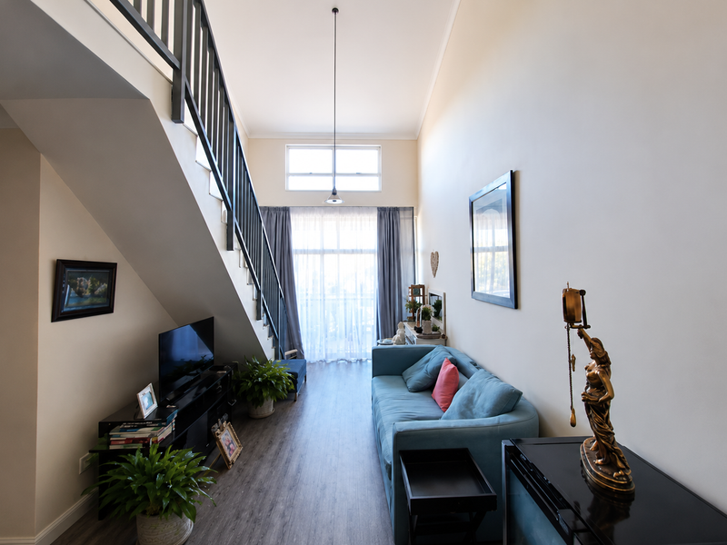 A bright living room featuring high ceilings, a staircase with a metal balustrade, laminate flooring, and large windows allowing for natural light.