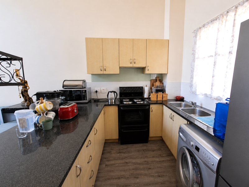 A functional kitchen featuring light wood cabinetry, granite countertops, a double sink, and a built-in washing machine with a modern laminate floor finish.