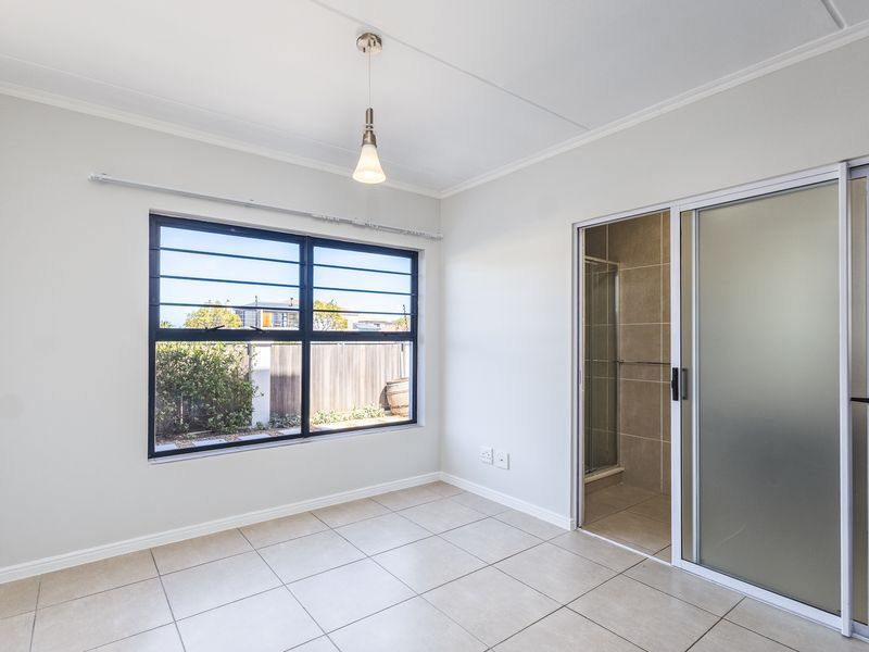 A bright, empty bedroom featuring neutral tiled flooring, a large window with security bars, and direct access to an en-suite bathroom via a sliding door.
