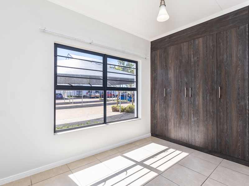 A bright, empty bedroom featuring modern dark wood-grain built-in cupboards, neutral tiled flooring, and a large window with security bars.